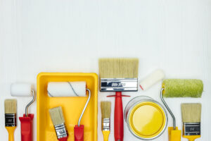 various paint brushes, rollers, paint cans and paint tray on white wooden background