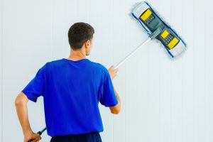 A young Caucasian man in a blue T-shirt washes the white wall of the room with a mop
