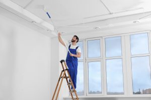 Handyman painting ceiling with roller on step ladder in room